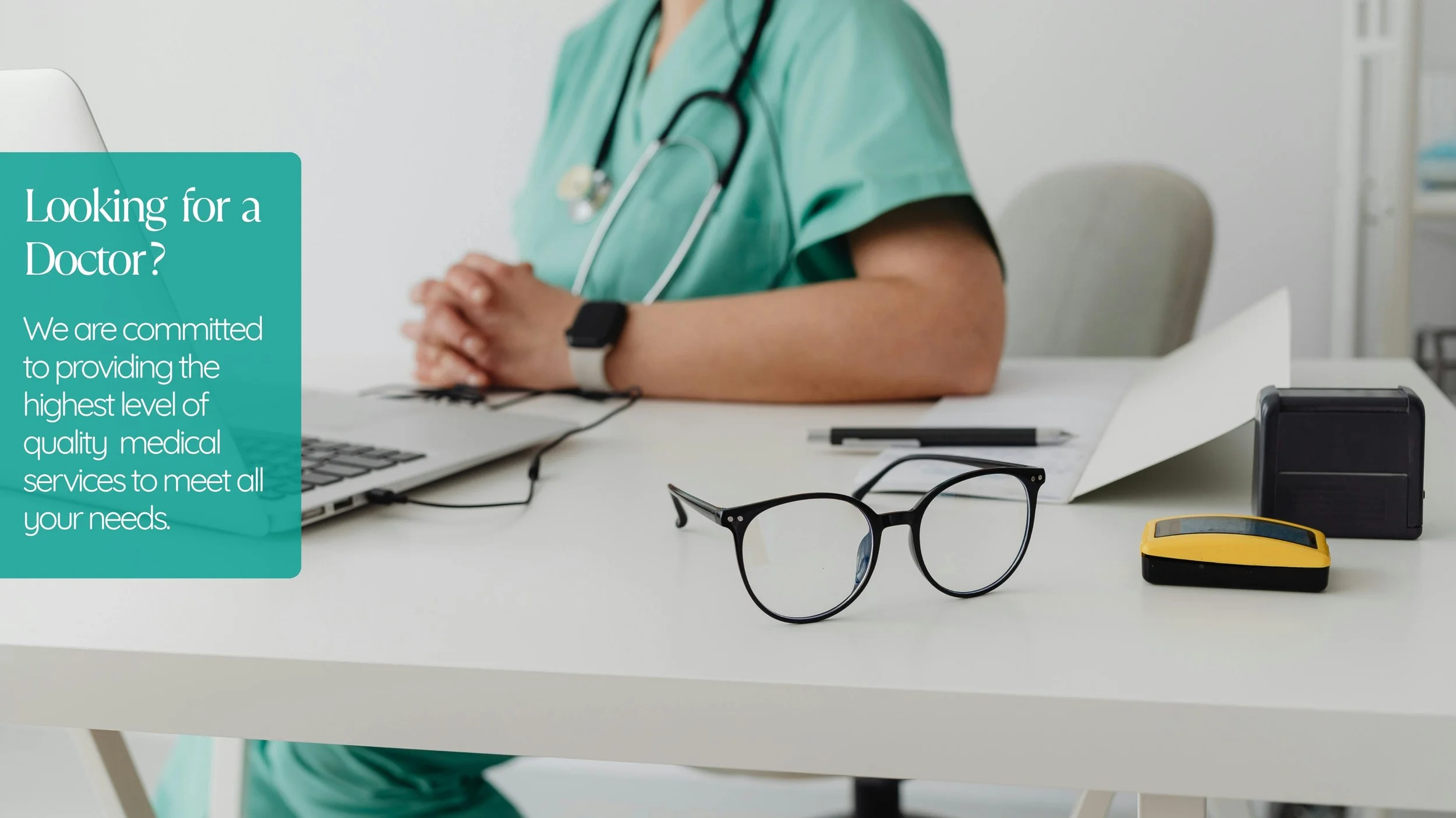 A medical professional in green scrubs sitting at a white desk, with a stethoscope around their neck, a laptop, a pair of glasses, and office supplies in front of them.