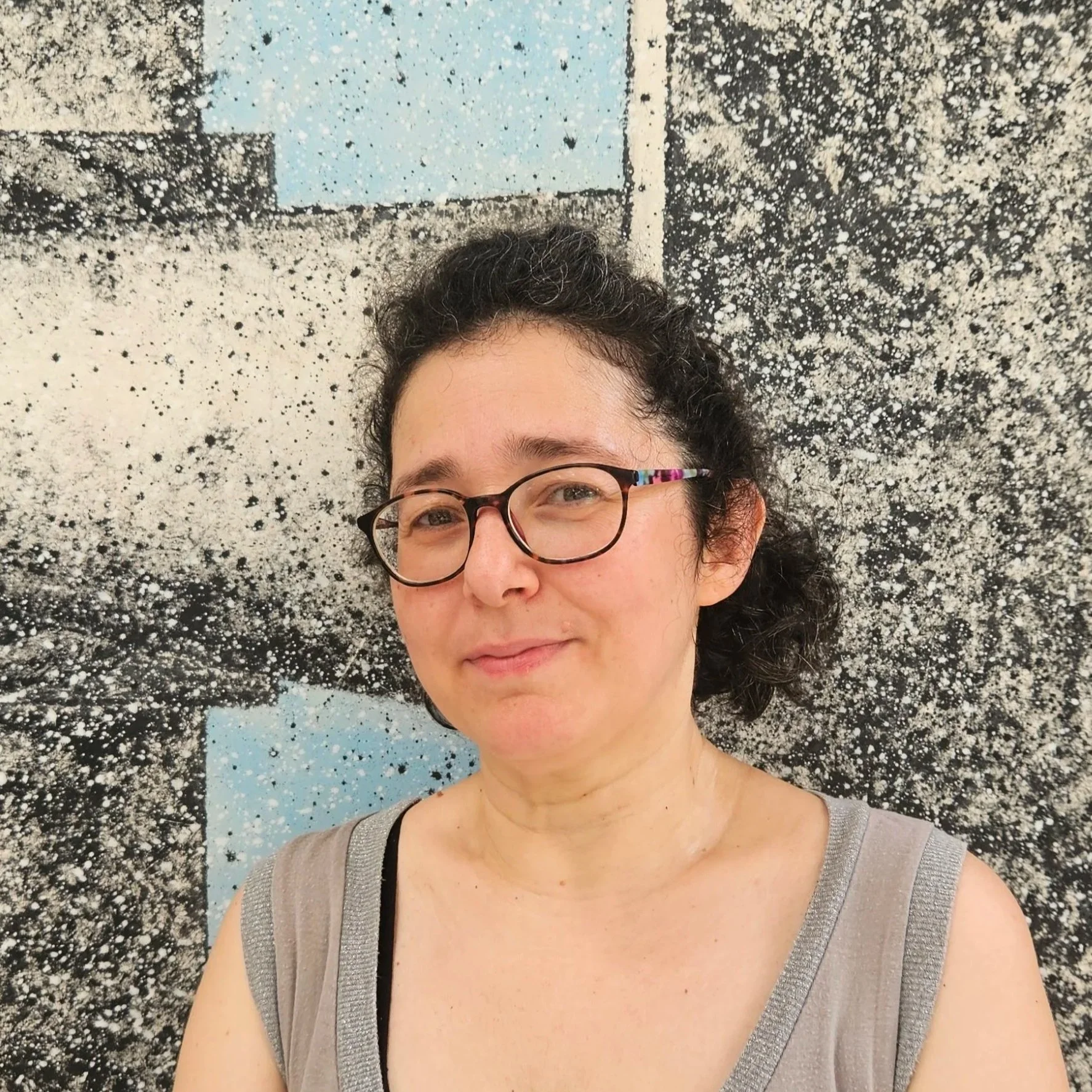 Primo piano di una donna con occhiali e capelli ricci scnografa di ATTOSECONDO, sorridente, davanti a uno sfondo di un'opera d'arte astratta con colori nero, bianco e azzurro.