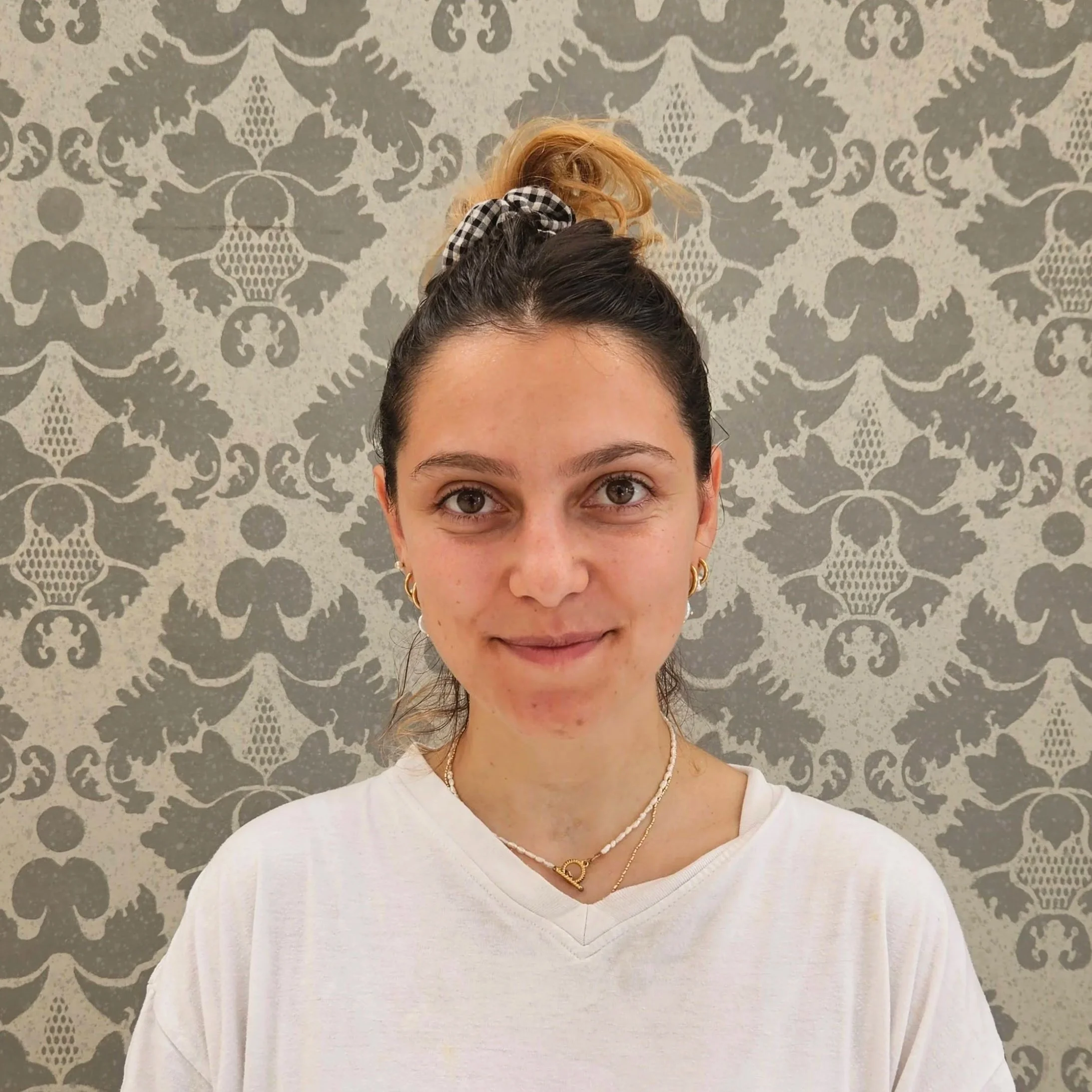 Donna scenografa di ATTOSECONDO Scenografie con i capelli raccolti, indossa una collana oro con una maglietta bianca, di fronte a una parete con motivo floreale grigio e bianco.