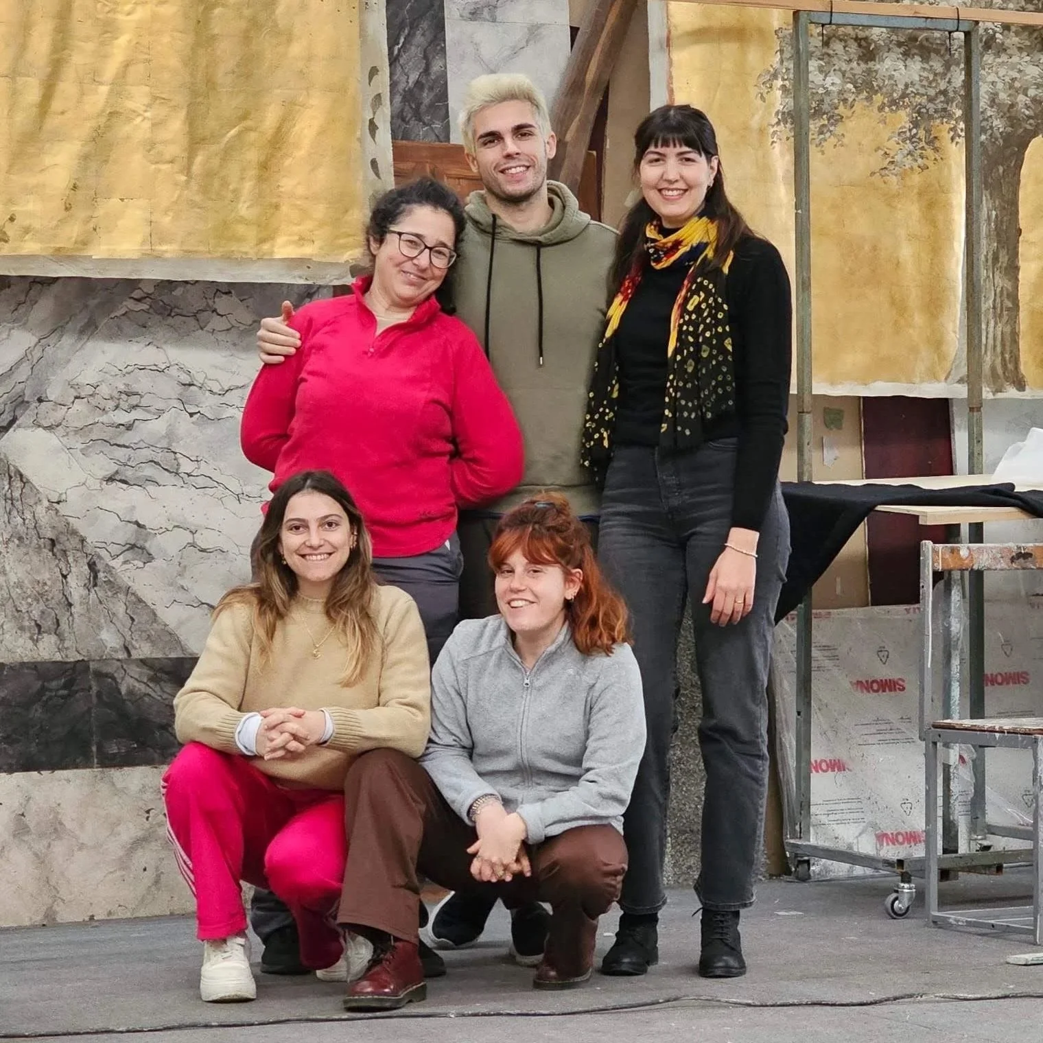 Un gruppo di cinque persone sorridenti e felici di ATTOSECONDO Scenografie posano insieme in un ambiente interno, tra cui un fondale di marmo e alcune attrezzature di scena, probabilmente in un teatro o uno studio di registrazione.