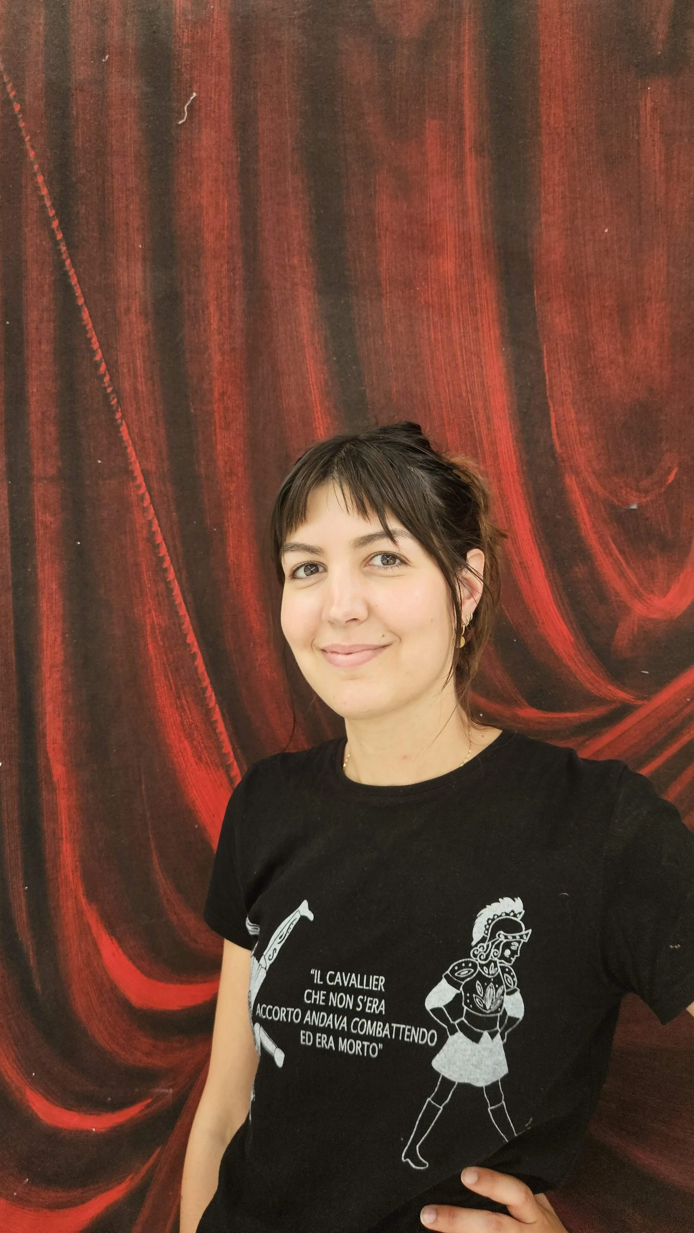 Donna presidente di ATTOSECONDO Scenografie con capelli corti e sorriso, indossa una maglietta nera con stampa, posizionata davanti a uno sfondo con un drappo rosso dipinto.