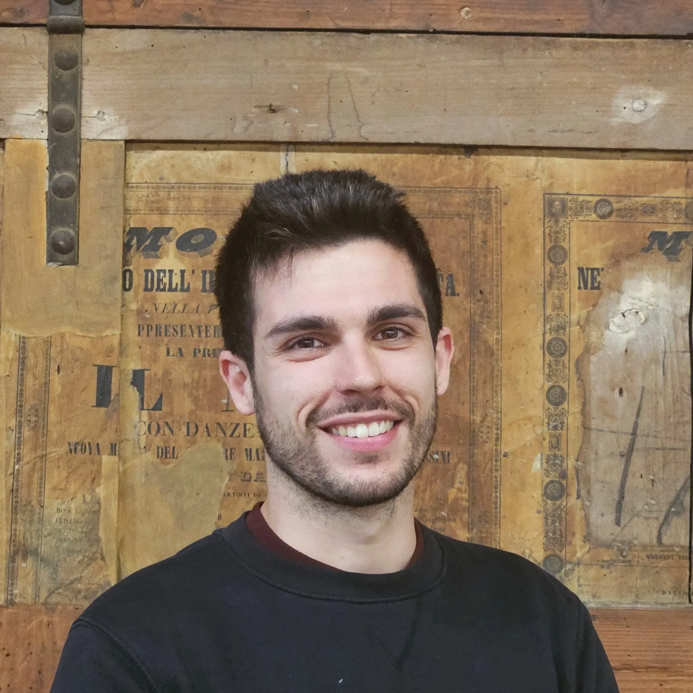 Primo piano di un uomo scenografo presso ATTOSECONDO Scenografie, con capelli castani scuri e occhi castani. Ha uno sfondo di pannelli di legno con antichi manifesti teatrali della sala di scenografia del Teatro Comunale di Modena.