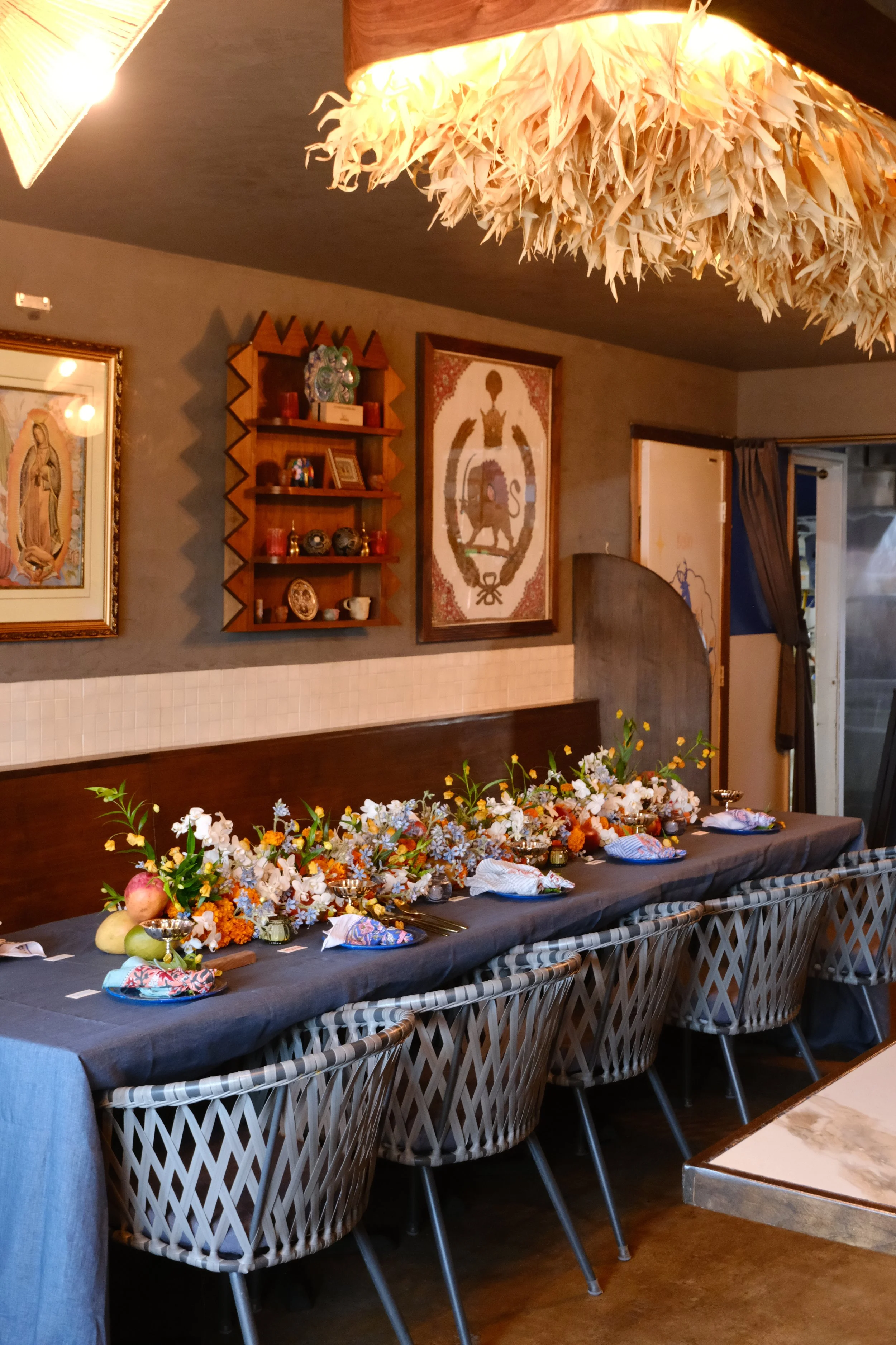 A long dining table covered with a dark tablecloth, decorated with a large floral centerpiece and place settings, situated in a cozy, warmly lit room with framed artwork and wall shelves.