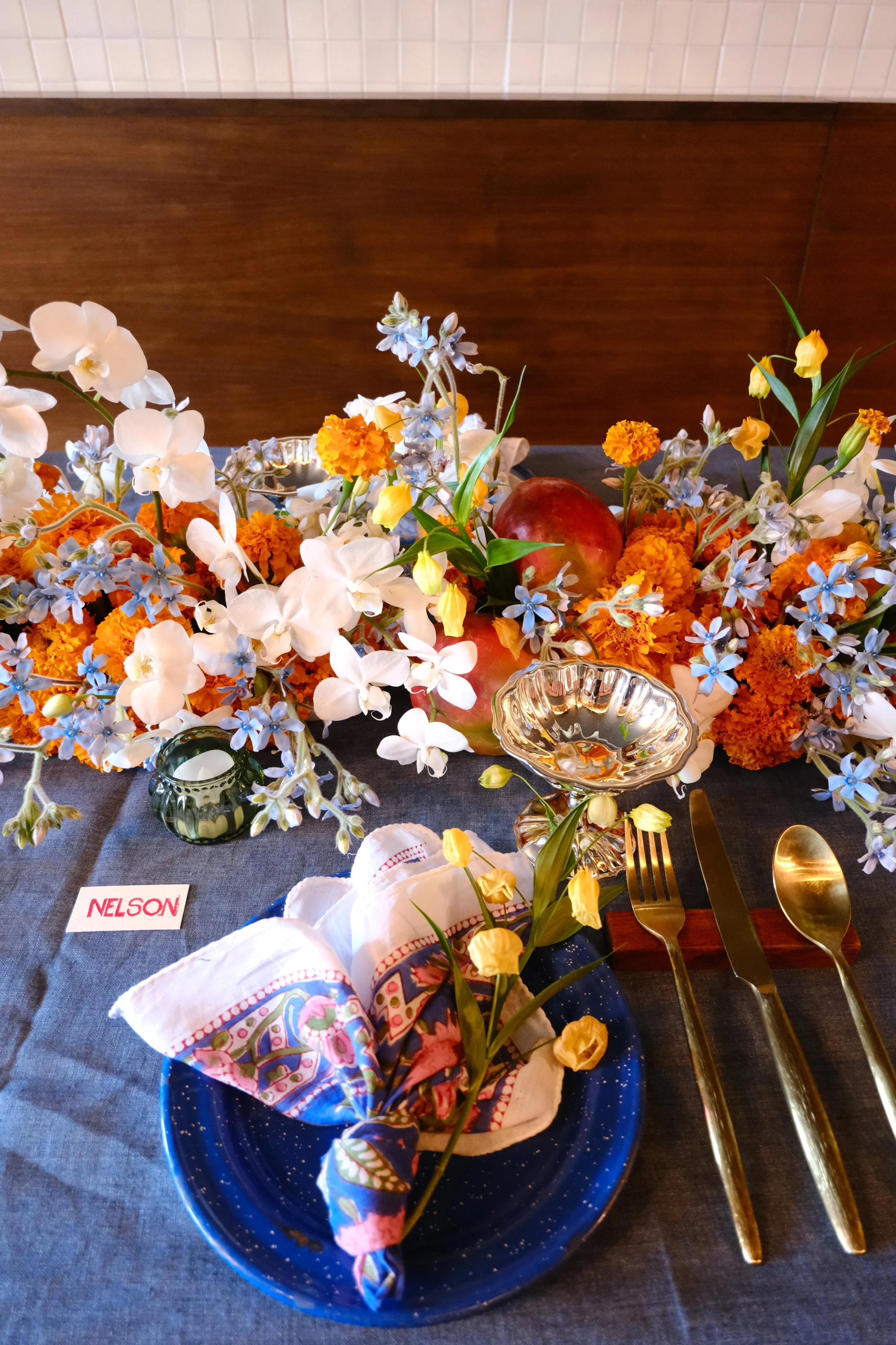 A table setting with a floral centerpiece, gold cutlery, a blue plate with a decorated napkin, and a small candle holder on a dark tablecloth. There is a name card labeled 'NELSON'.