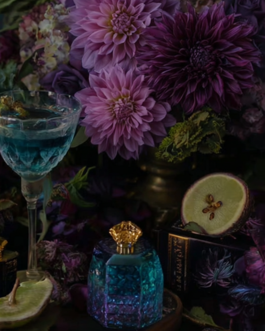 Still life featuring pink and purple dahlias, a glass of blue liquid with floral garnish, lemon slices, and a vintage perfume bottle - symbolising sensory richness and grounded femininity.