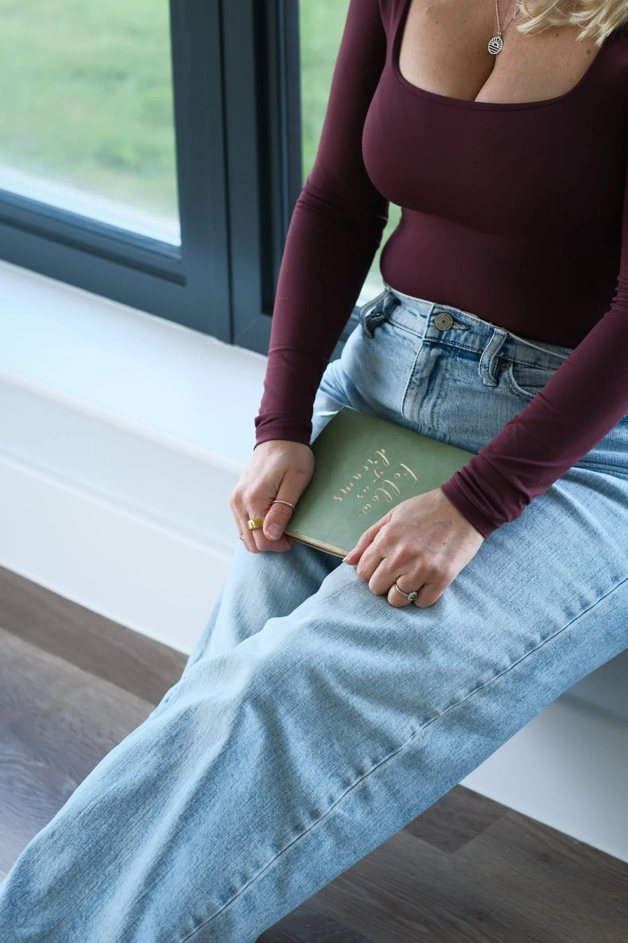 Woman sitting on a windowsill holding a book about calm and relaxation, representing nervous system regulation, self-awareness and intentional rest for high-achieving women