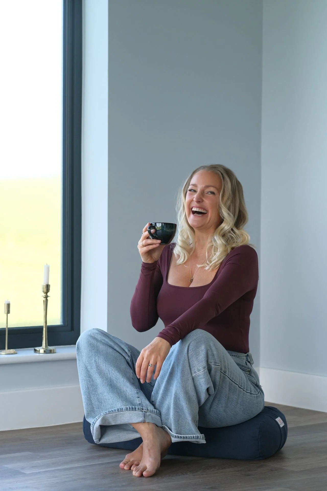 Woman sitting on a floor cushion with coffee near a window and candles, representing slow mornings, nervous system regulation and intentional living for women