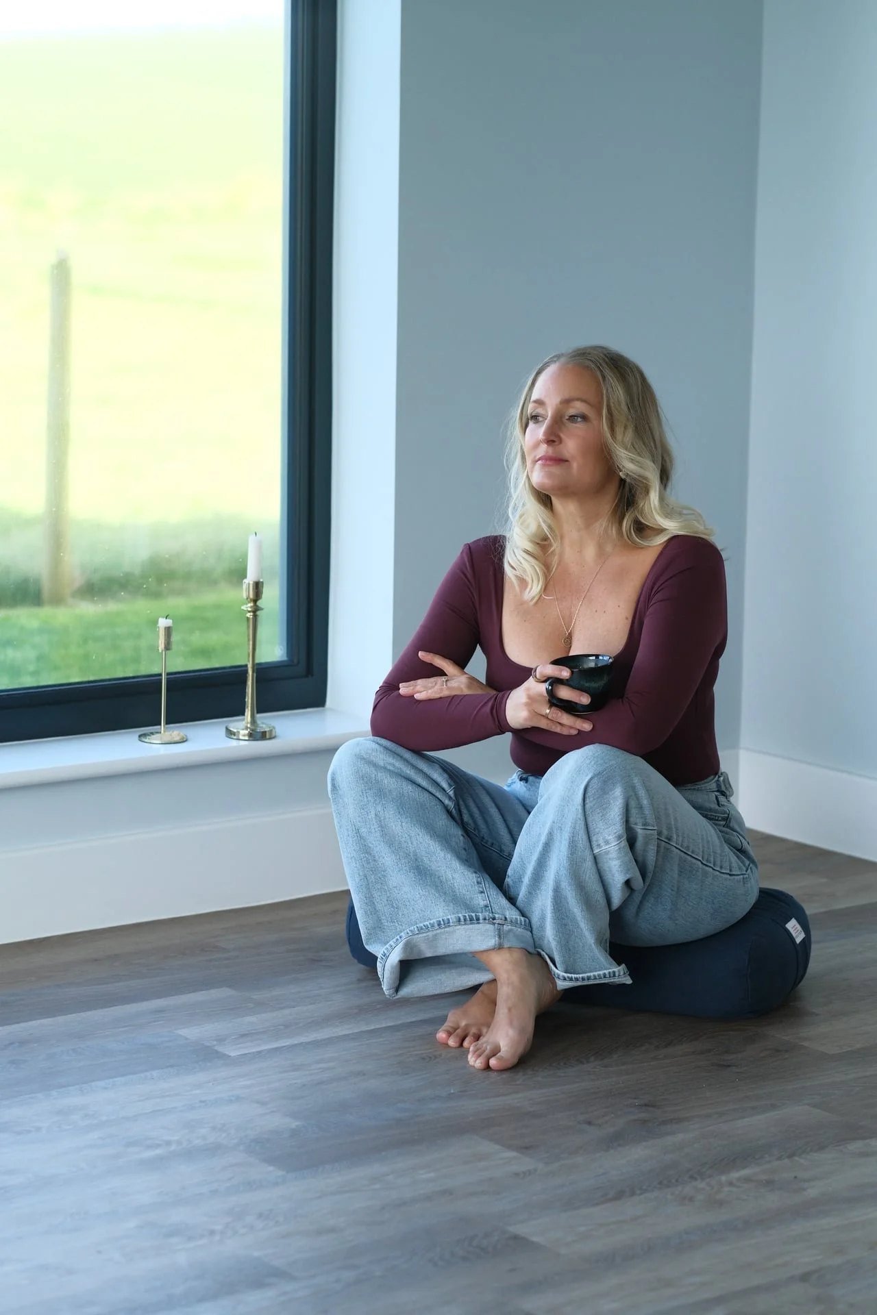 Woman sitting cross-legged by a window holding a coffee mug in a calm home setting, representing reflection, nervous system regulation and intentional living for women