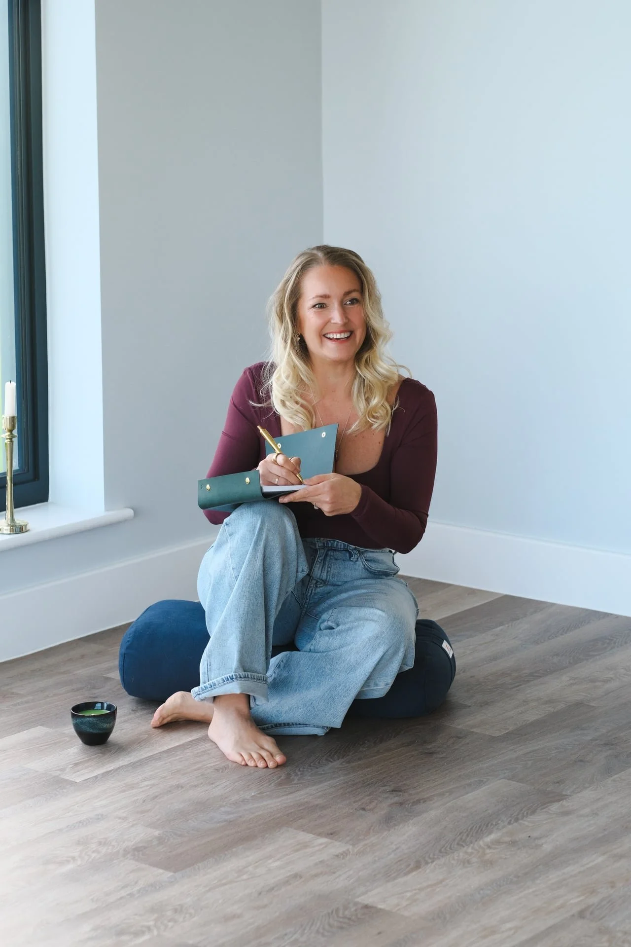 Woman sitting cross-legged journaling by a window in a calm home setting, representing reflection, clarity and nervous system awareness in somatic coaching work