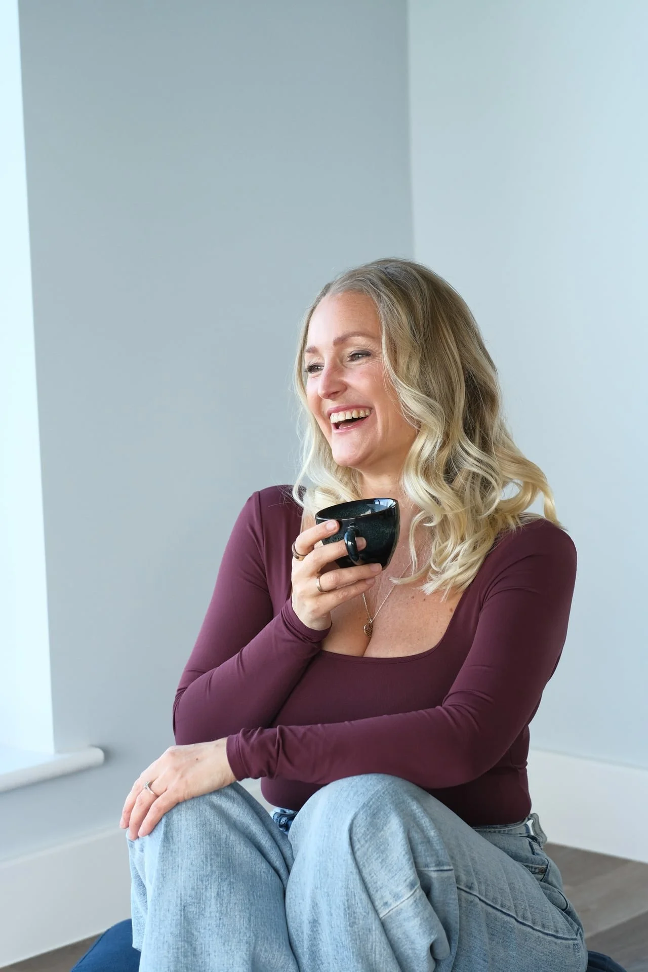 Woman sitting with a coffee mug in a calm, light-filled space, representing ease, presence and nervous system regulation for high-achieving women