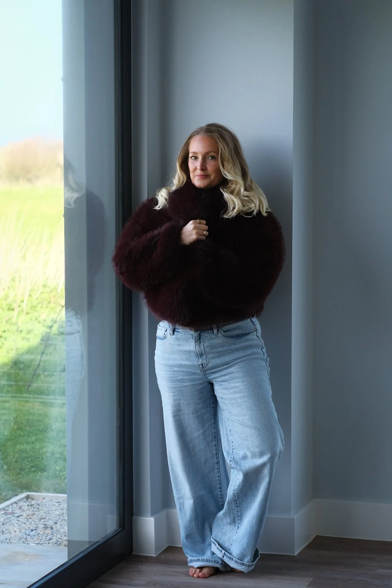 Woman standing barefoot by a large window in a calm home setting, representing nervous system regulation, presence and connection to self