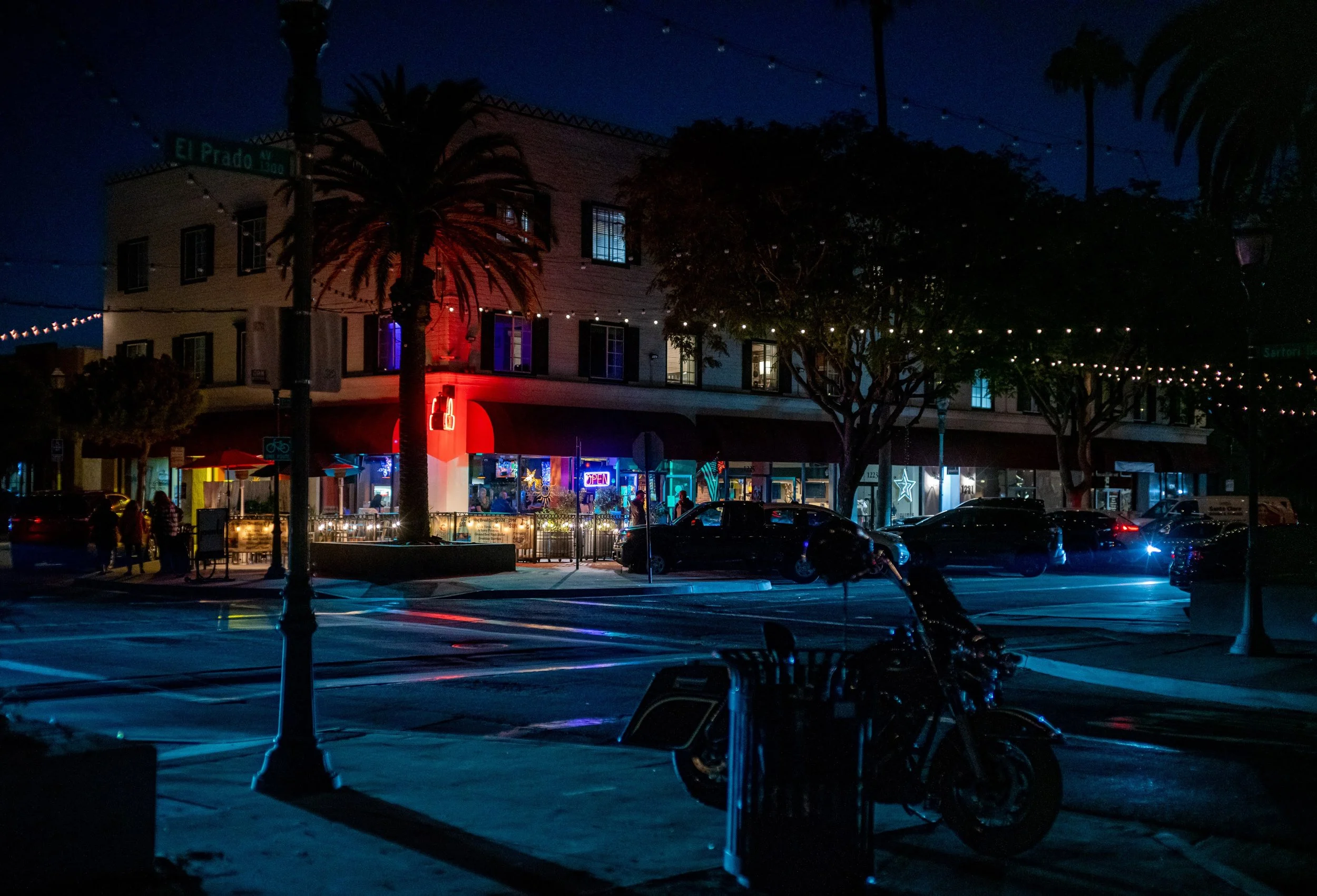 Nighttime street scene with a building lit up with colorful neon signs, cars parked in front, trees with string lights, and a motorcycle at a bus stop.