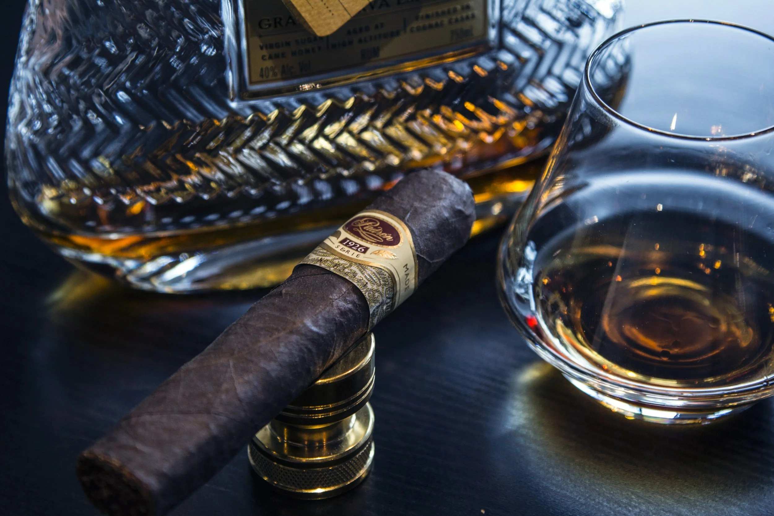 A glass of whiskey, a cigar resting on a small gold-colored stand, a bottle of whiskey with a textured glass surface, and a whiskey glass all placed on a dark surface.
