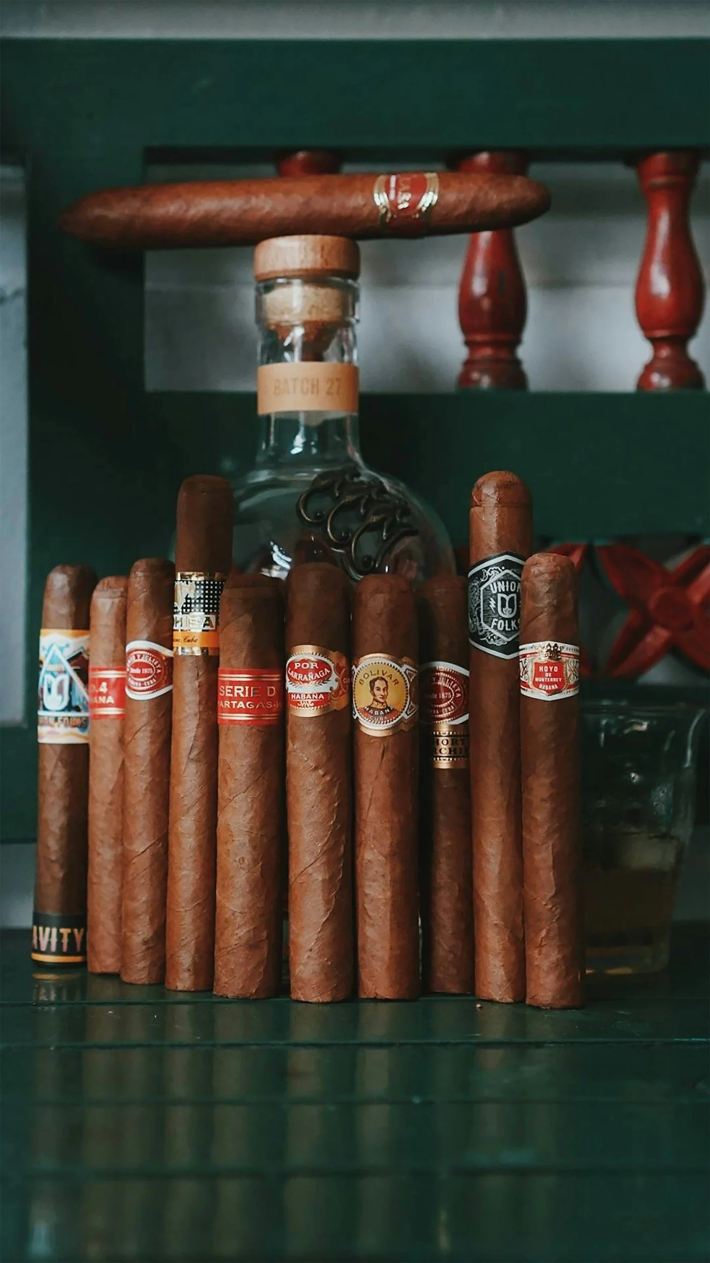 A collection of cigars arranged in front of a glass bottle with a cork, on a dark green wooden surface.