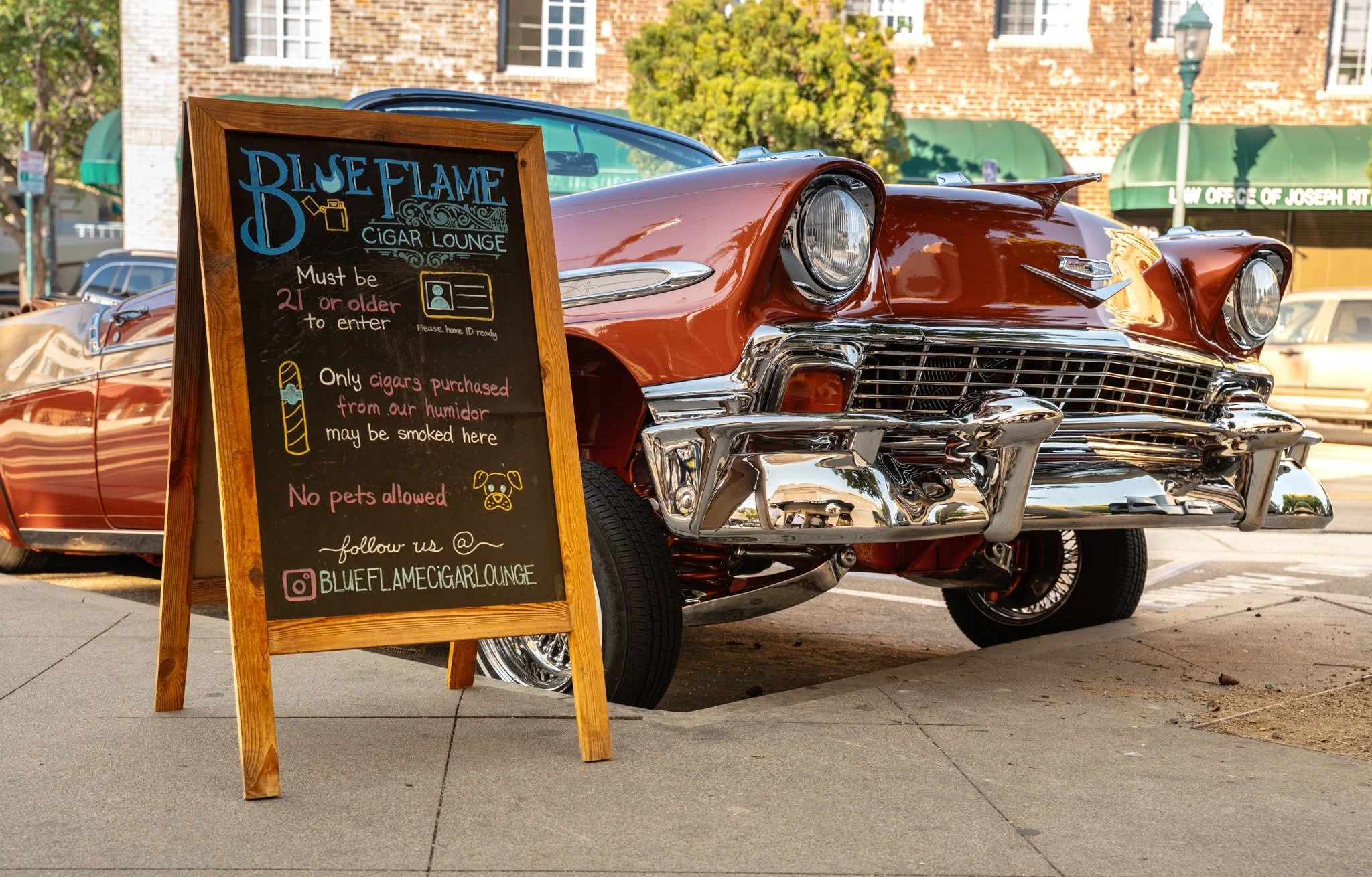 A vintage orange car parked on a sidewalk with a blackboard sign in front of it. The sign indicates the location is the Blue Flame Cigar Lounge, with rules about age, smoking, and pets, and a social media handle for the lounge.