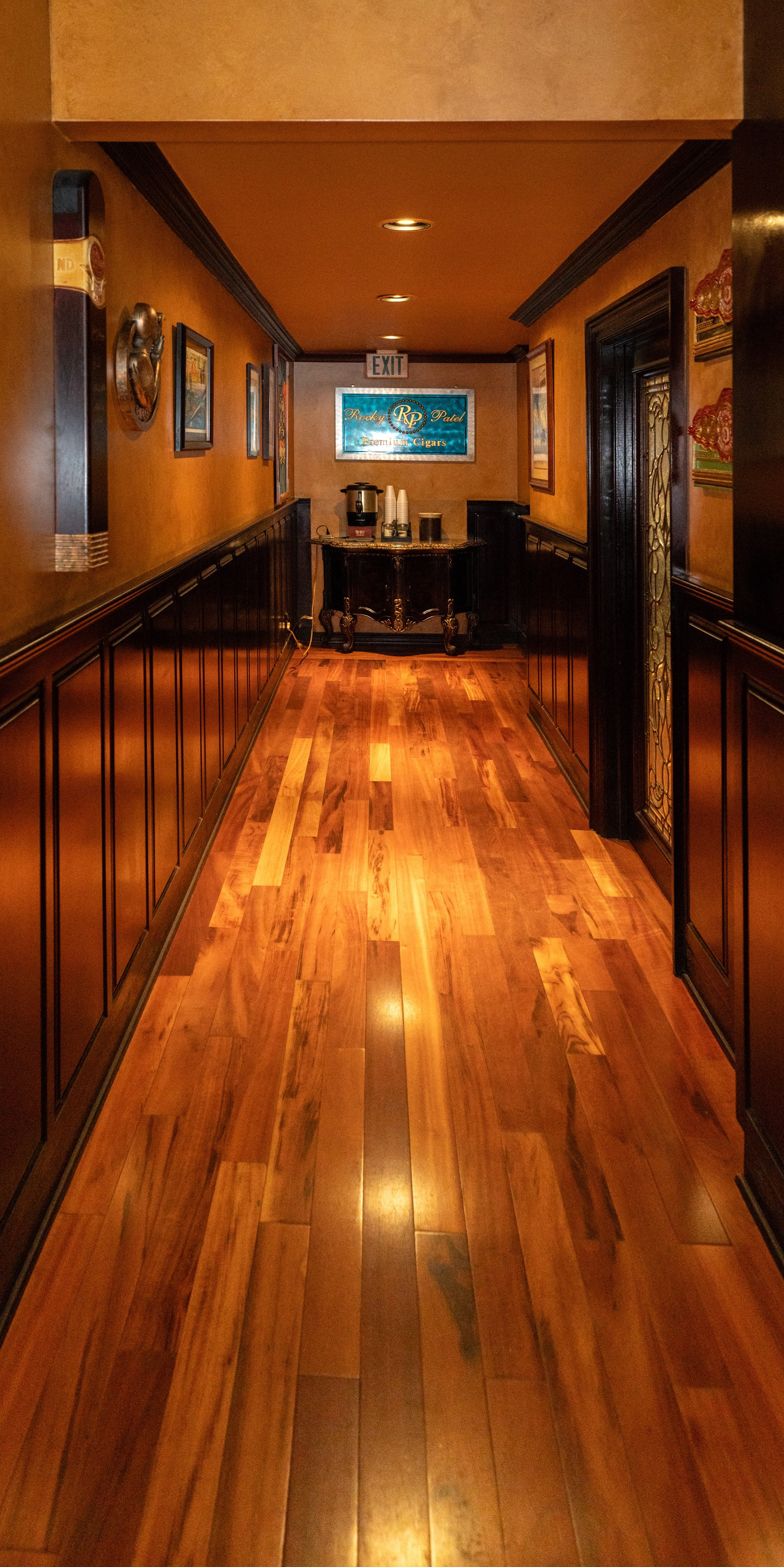 A corridor with wooden flooring, framed pictures on the walls, and a small table with a coffee urn and cups at the end. Sign indicating 'EXIT' above the table.