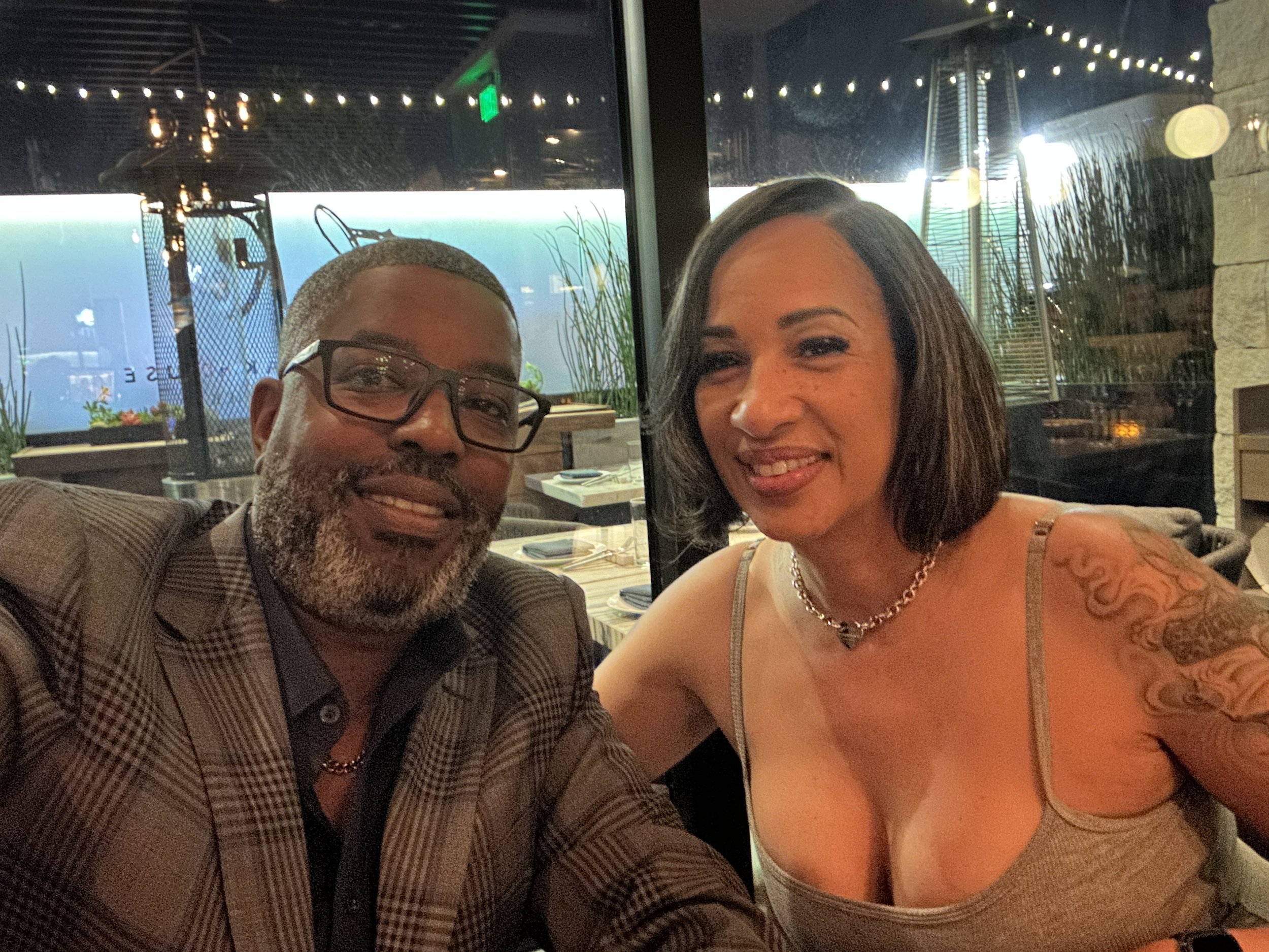 Lyman and Nacy smiling at a restaurant table, with a large window showing an outdoor patio area with string lights and plants.