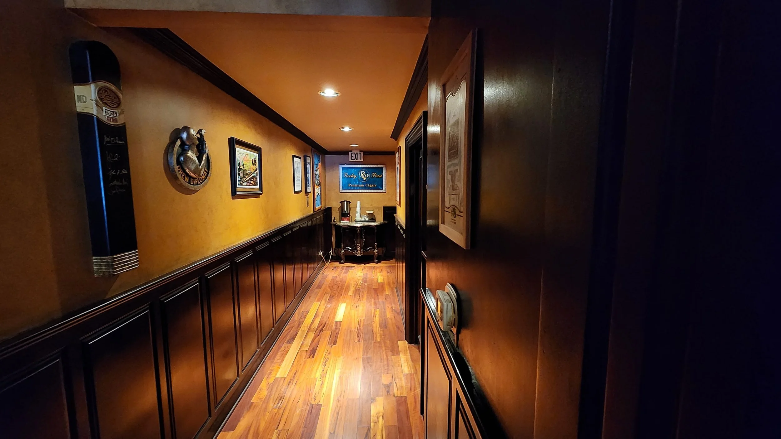 A narrow hallway with wooden paneling and a yellow-orange textured wall, decorated with framed pictures and a decorative plaque, leading to a small table with a coffee maker and cups, with an exit sign and a blue sign that reads "Premium Cigars" at the end.