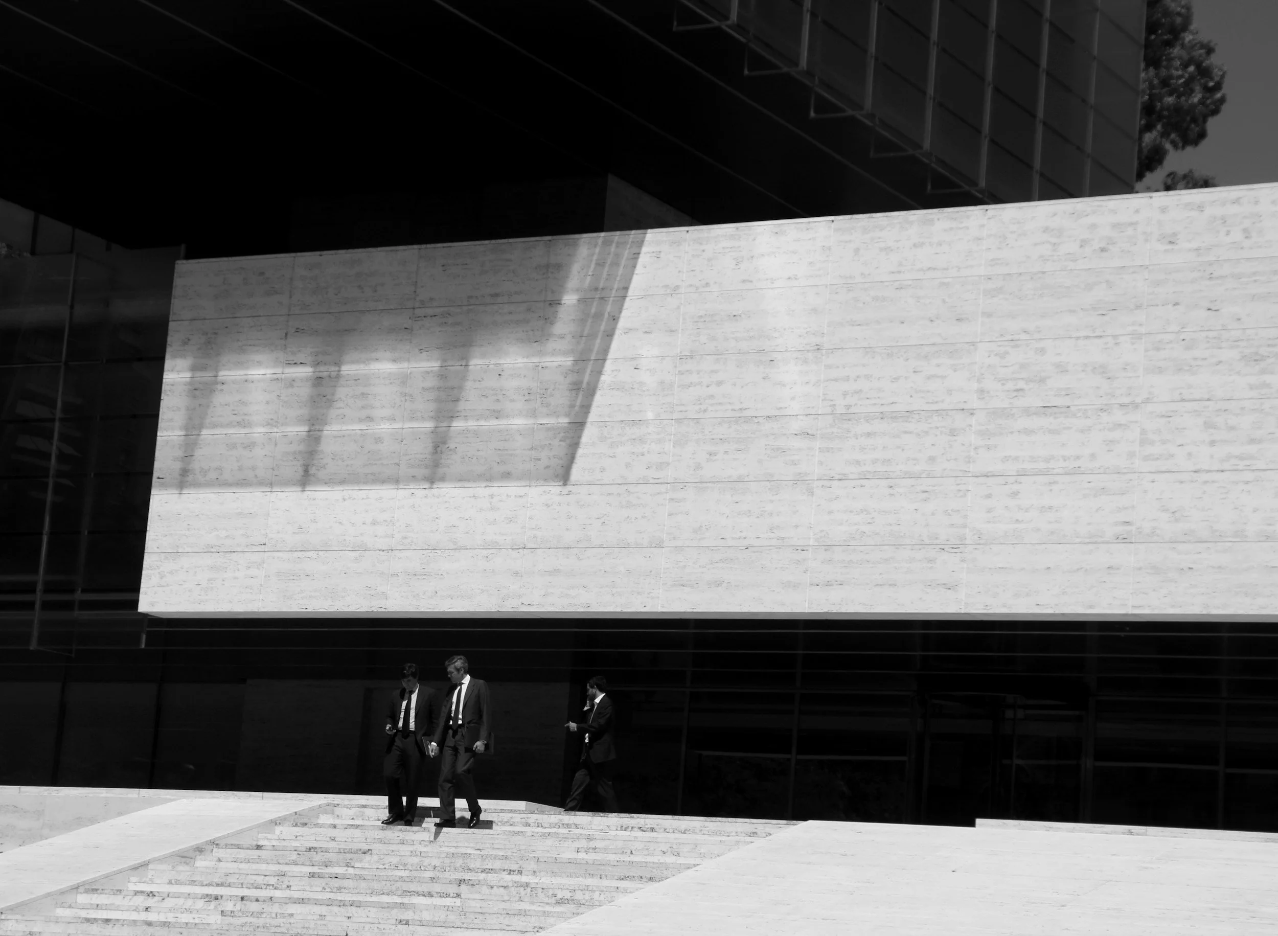 Three men in business suits walking down steps outside a modern building with large, textured, white facade and dark glass windows, shadows cast on the wall.