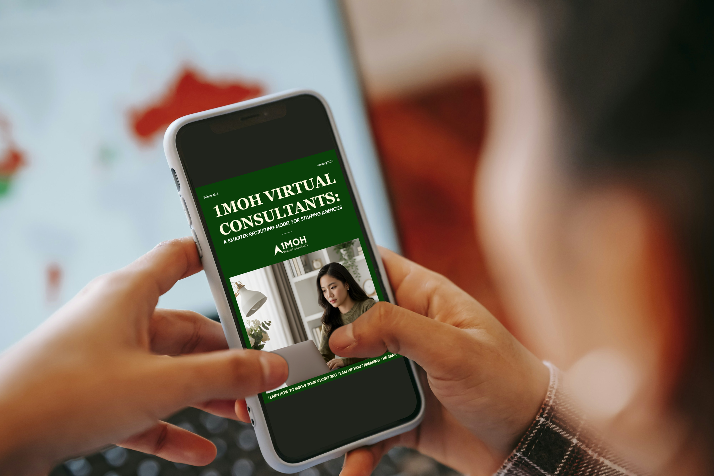 Person holding a smartphone displaying a digital magazine titled '1MOH Virtual Consultants,' with a woman working on a laptop on the cover.