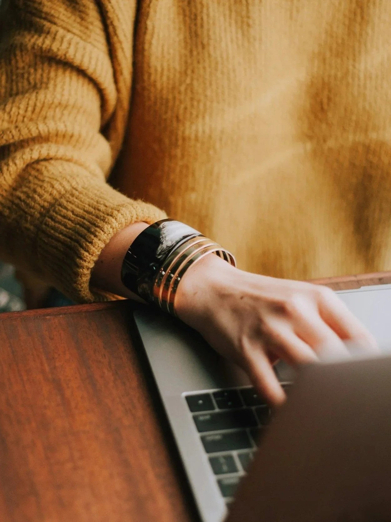 Close-up of a person's hand using a laptop, wearing a layered silver and black bracelet, and dressed in a mustard-colored sweater.