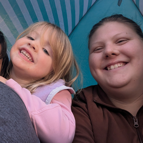 Two smiling girls, one with blonde hair and one with darker hair, lying inside a tent during daytime.