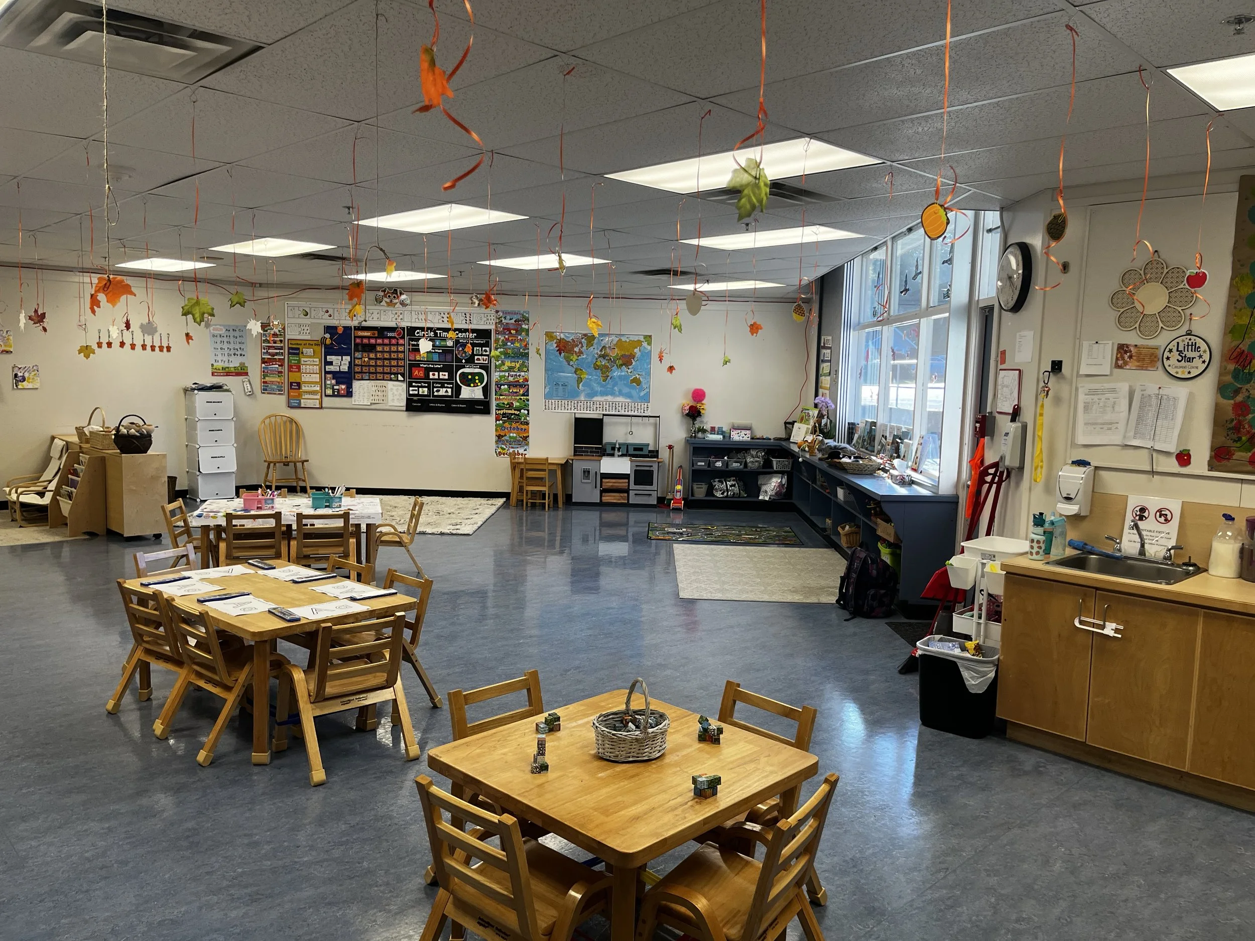A classroom with small tables and chairs, decorated for fall with hanging orange and green leaves and ribbons. There are shelves, a world map, and educational posters on the walls, with large windows letting in natural light.