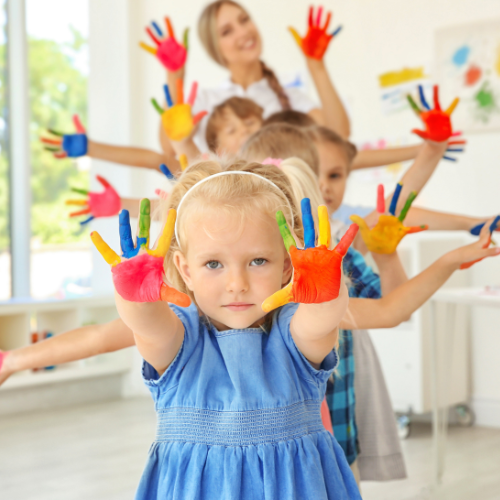 Children with painted hands raised in a room with sunlight, led by a smiling teacher in the background.