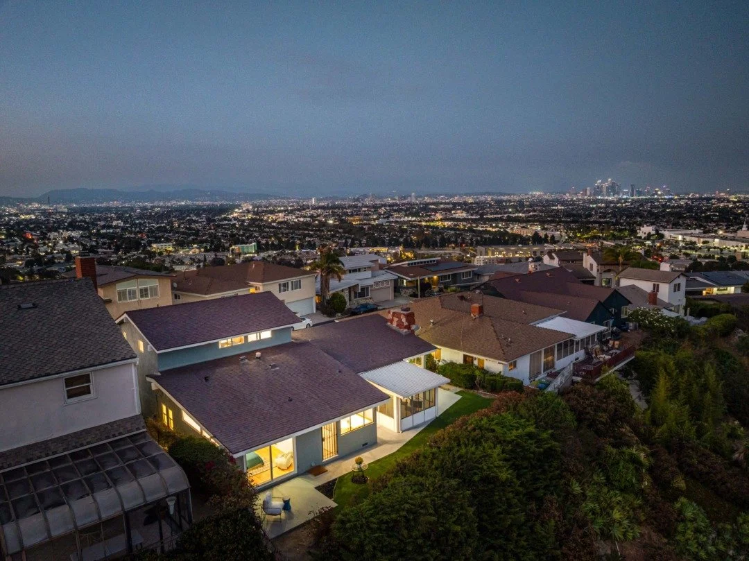 Nighttime aerial view of a suburban neighborhood with houses, some with outdoor lighting, overlooking a thriving cityscape with tall buildings and city lights in the distance.