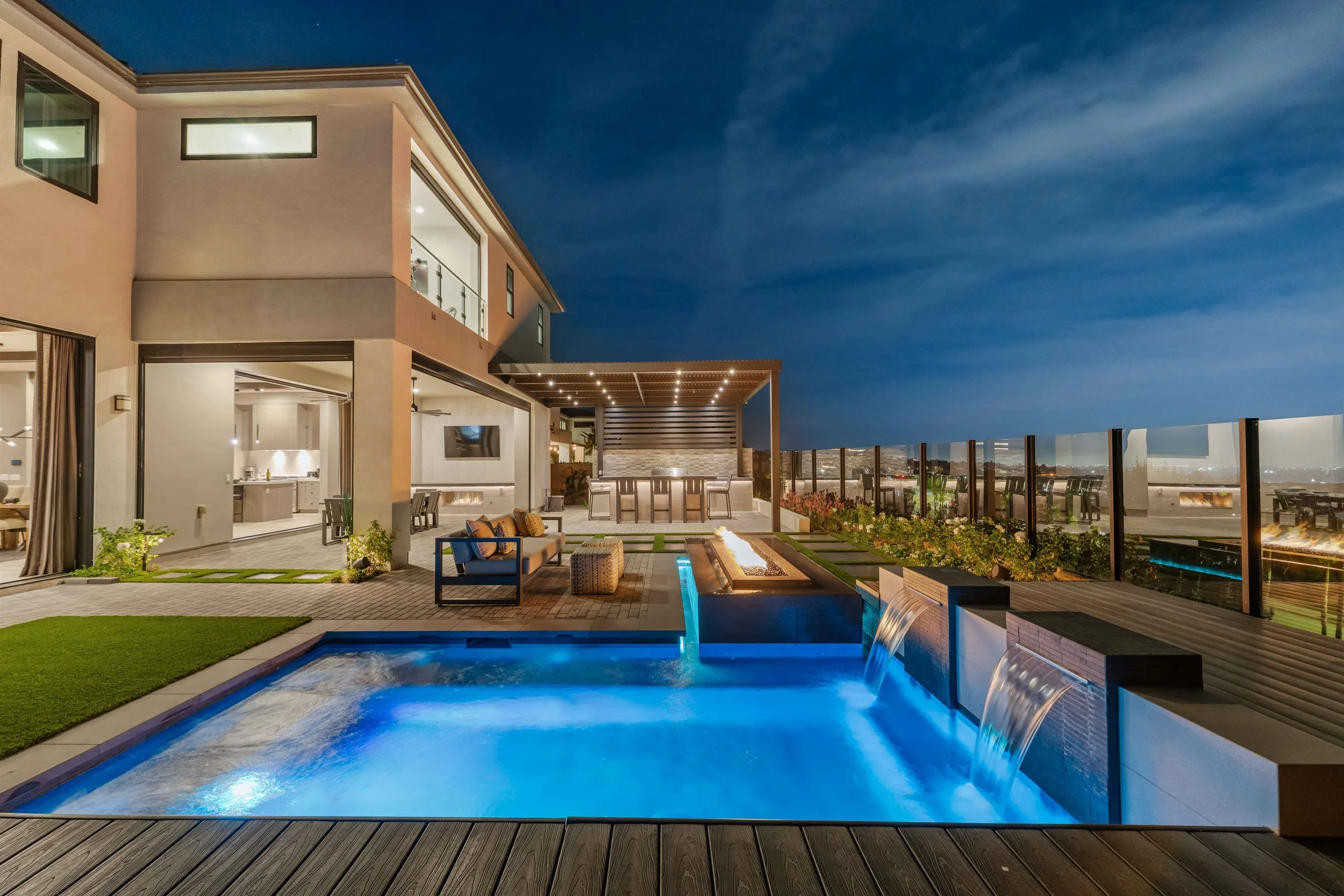 Night view of a modern house rooftop with a lit swimming pool, outdoor bar, seating area, and city lights in the background.
