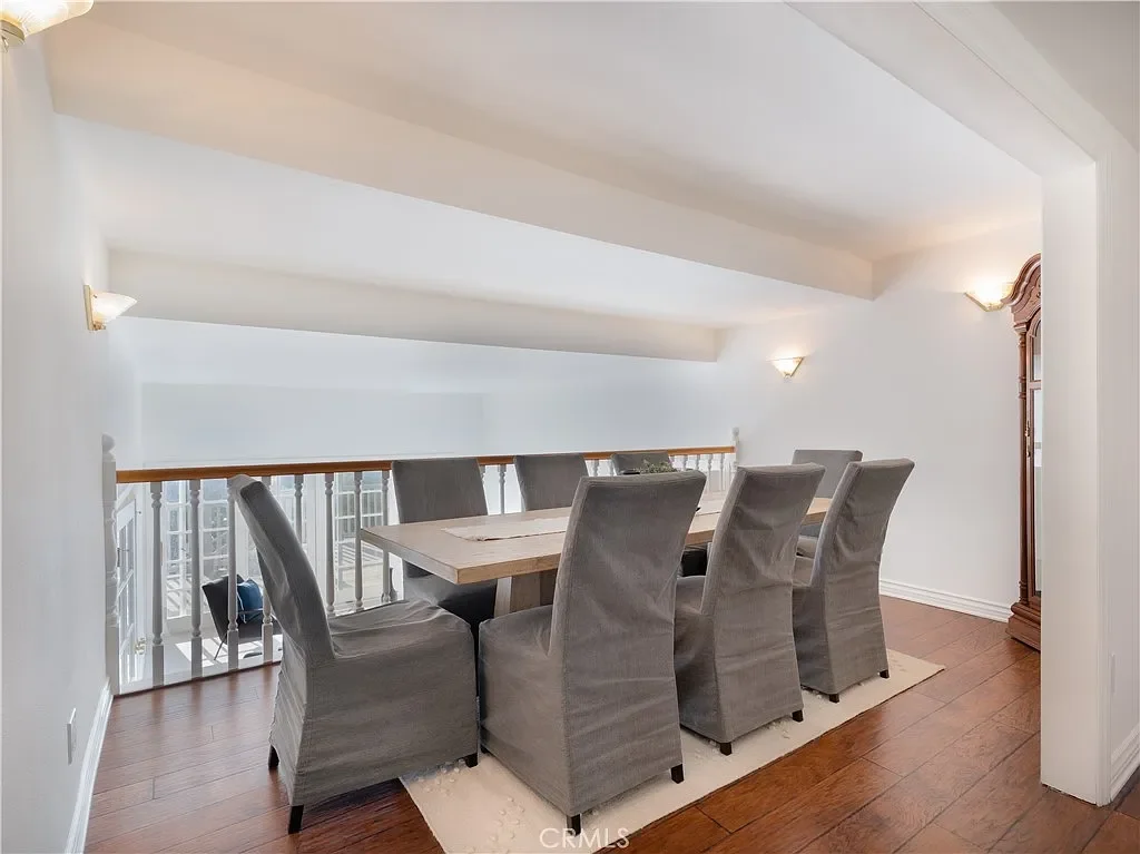 Dining area with eight upholstered gray chairs around a rectangular wooden table, hardwood flooring, and wall-mounted light fixtures, with a wooden grandfather clock in the corner.