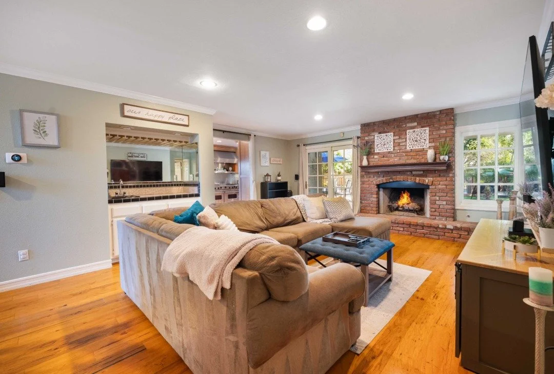 A living room with a brown sectional sofa, throw pillows, and a blanket, facing a brick fireplace with a lit fire. The room features hardwood floors, framed artwork, a television, and a cabinet. There are large windows and a sliding glass door lettin