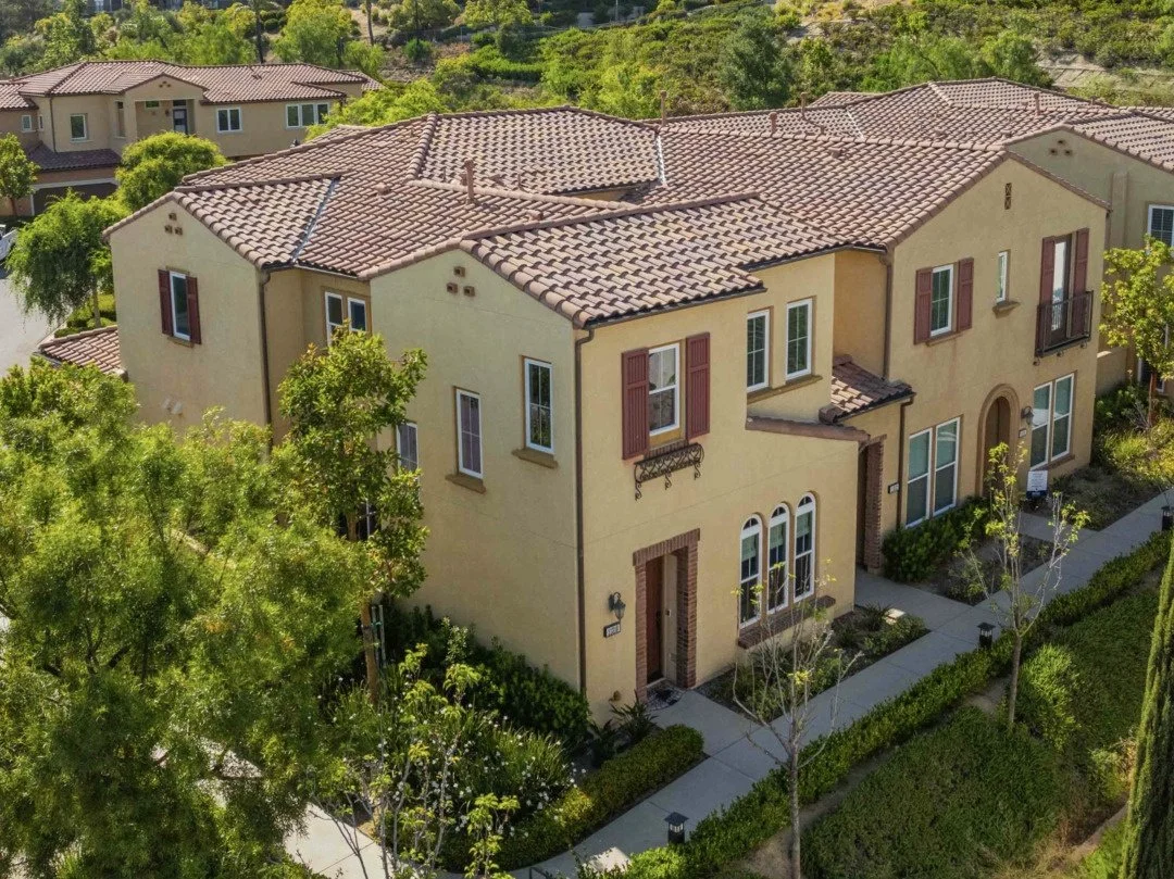 A two-story yellow stucco house with a red-tiled roof, surrounded by trees and landscaping, in a suburban neighborhood.