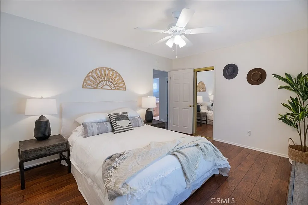 Bedroom with a white bed, gray and striped pillows, two black nightstands with lamps, ceiling fan, decorative wall art above bed, open closet door, doorway to another room, and a large green potted plant.
