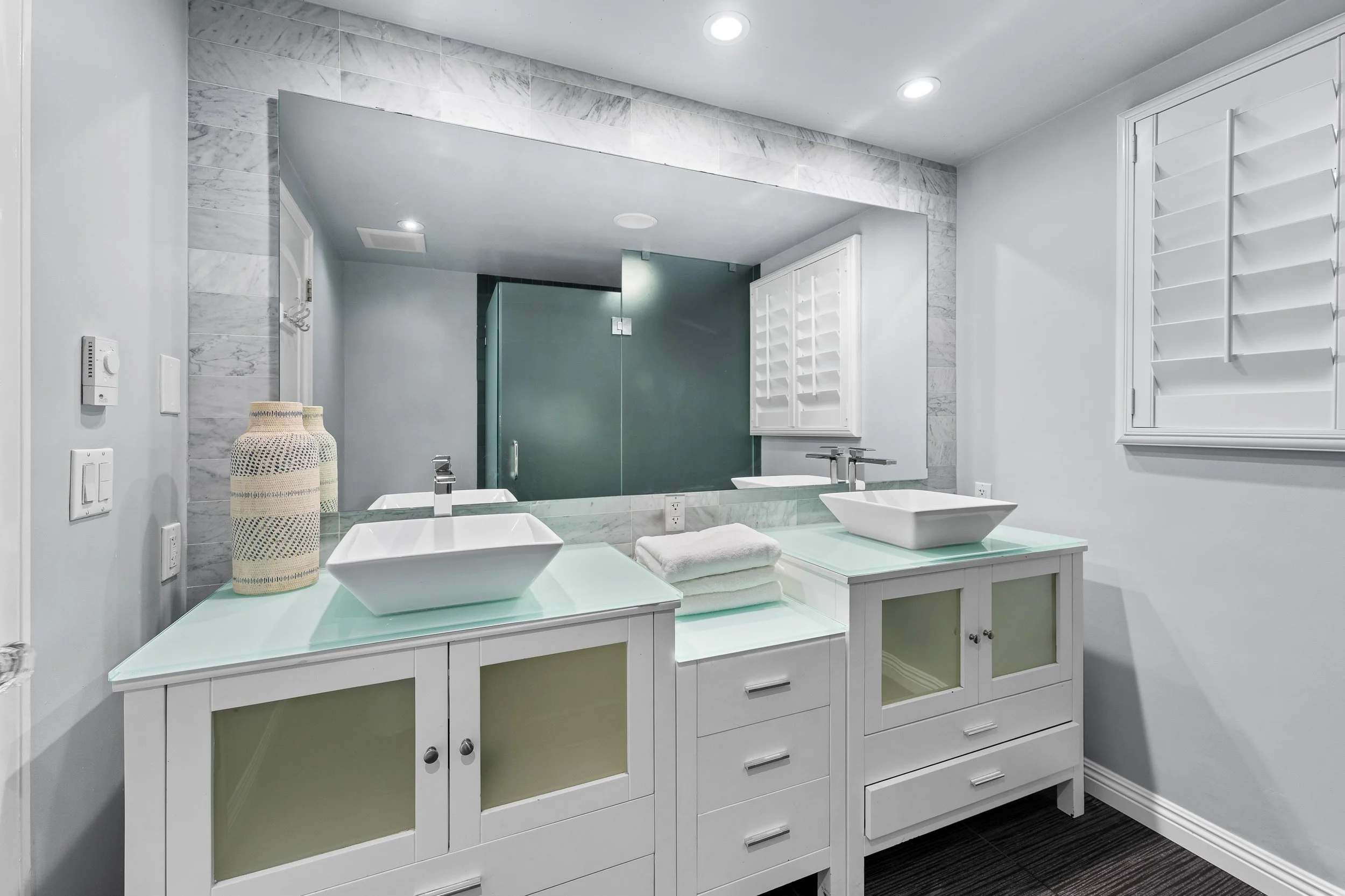 Modern bathroom with double vanity sinks, large mirror, white cabinetry, towel, decorative vases, and a glass shower door.