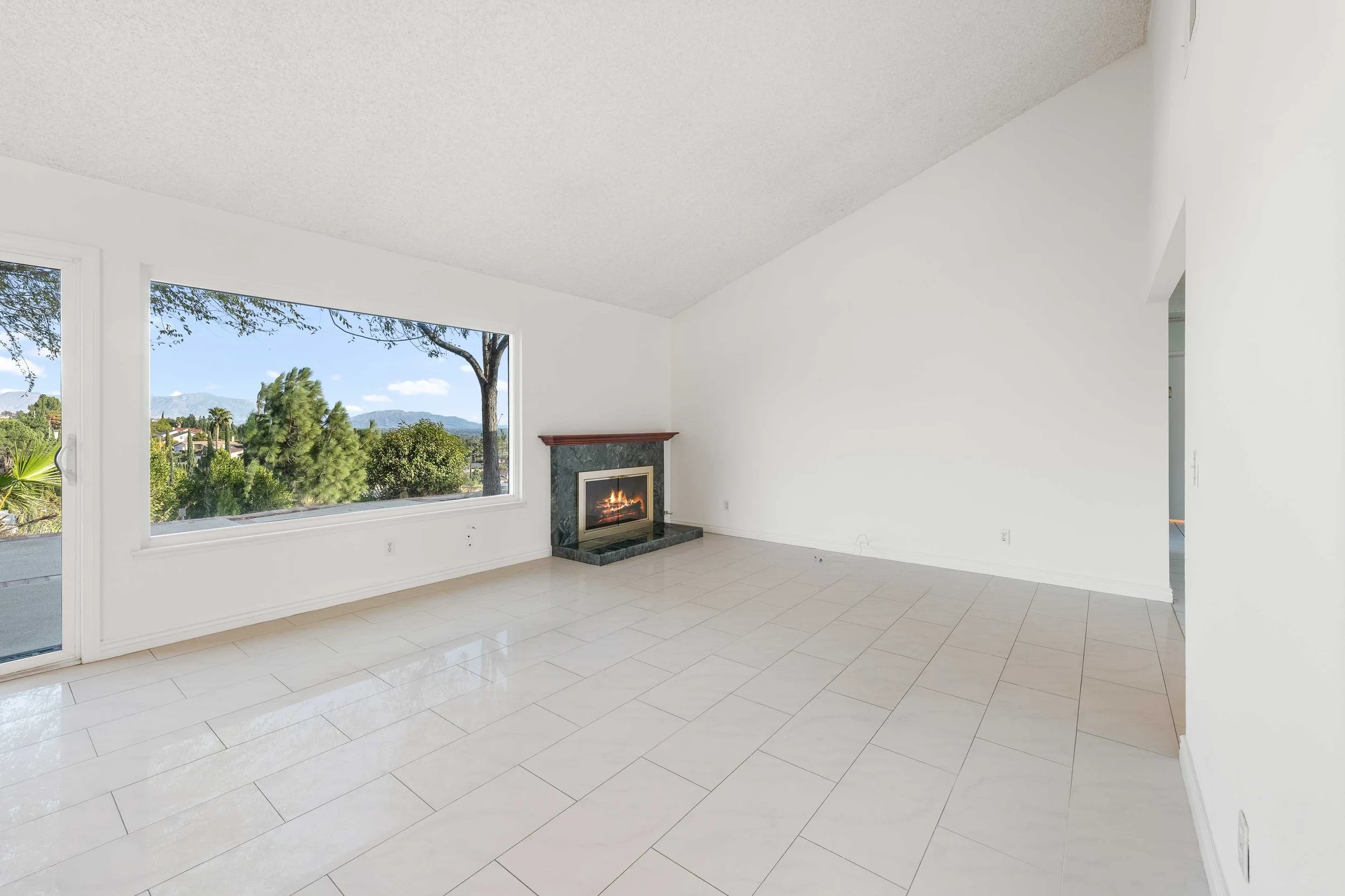 Empty living room with large window, tile floor, and a fireplace with flames, overlooking trees and mountains outside.