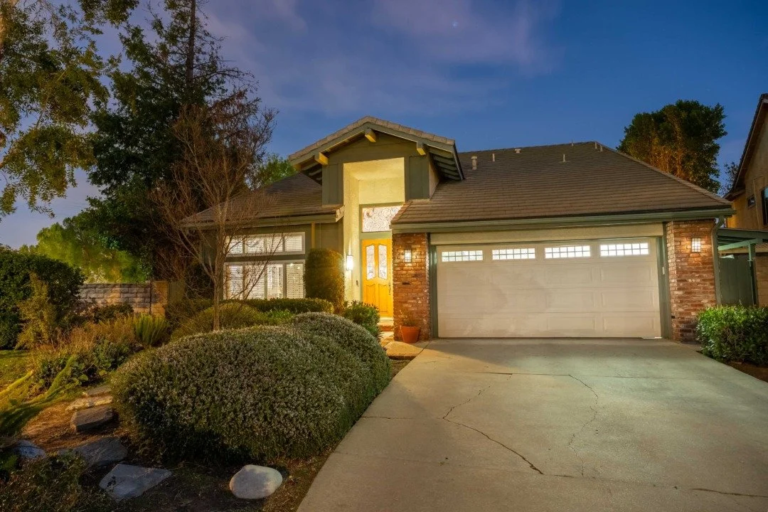 Exterior view of a house with a concrete driveway, brick and siding exterior, front door with glass panels, attached garage with small windows, landscaped front yard, and trees, during twilight.