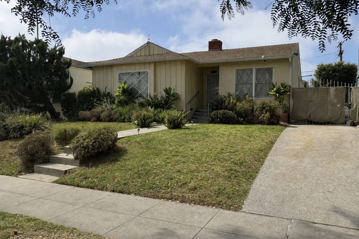 A yellow single-story house with a brown shingle roof, surrounded by green plants and shrubs. There are stairs leading up to the front door, and a driveway on the right side. The front yard has a small lawn and a concrete sidewalk.