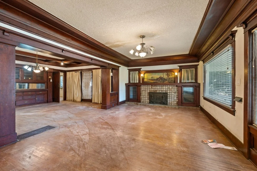 Empty living room with wooden trim, large windows with blinds, a fireplace, and built-in shelves and cabinets with glass doors.