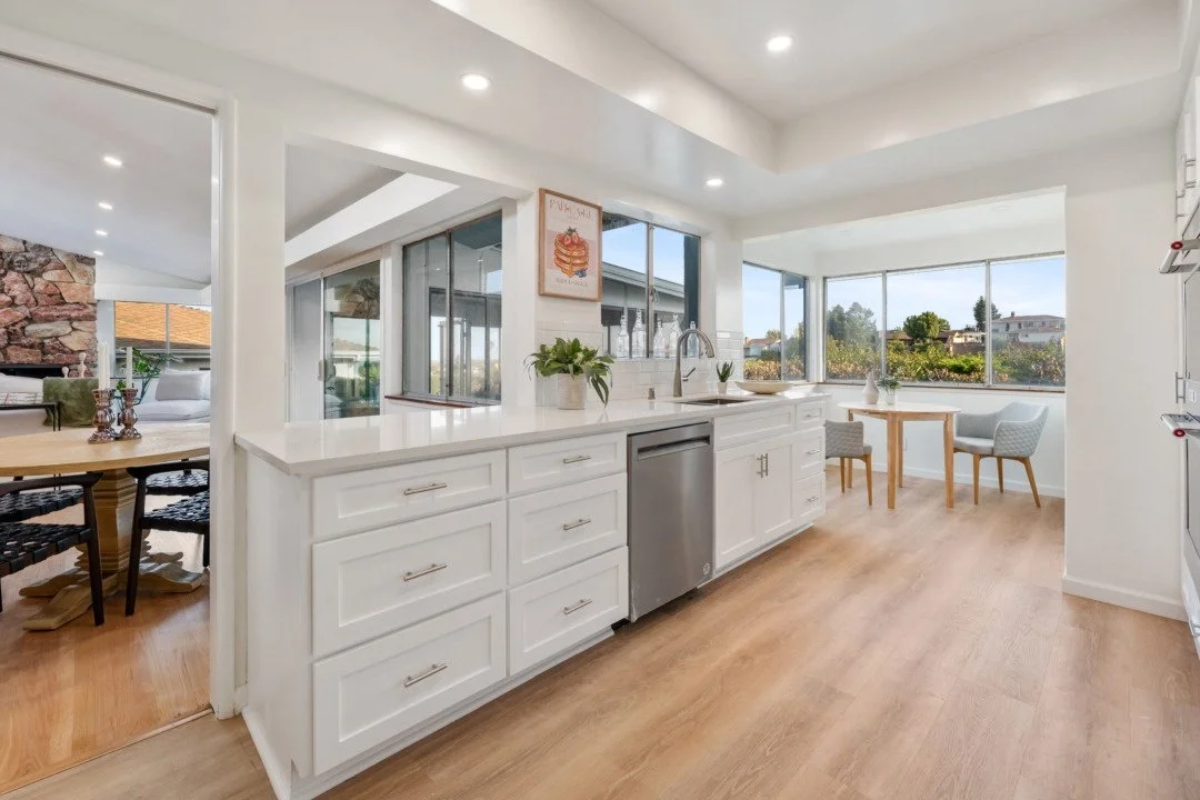 Bright modern kitchen with white cabinets, a small stainless steel dishwasher, and large windows overlooking an outdoor area with trees.