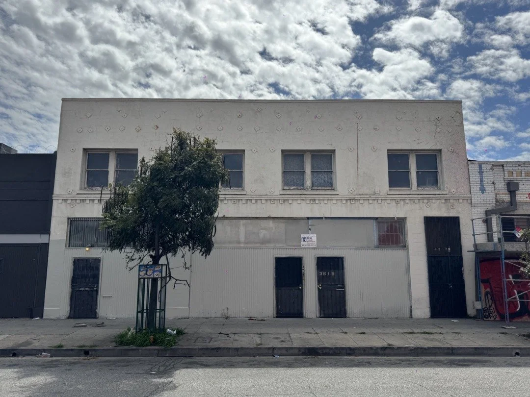 A two-story white building with four windows on the second floor, three of which are broken or missing glass. The first floor has two black doors and a small window, with a 