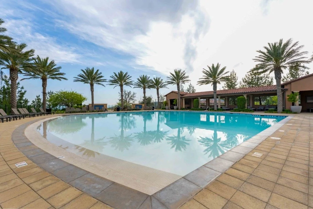 A luxurious outdoor swimming pool surrounded by palm trees and lounge chairs, with a building and cloudy sky in the background.