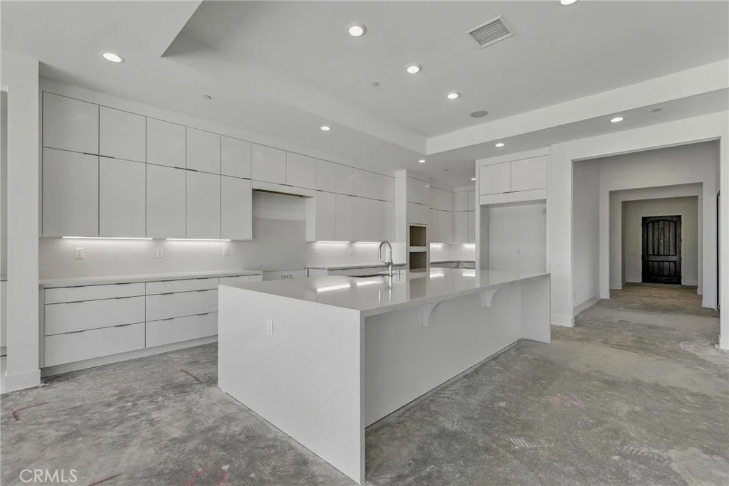 Modern white kitchen with minimalist cabinets, a large island with a sink, and no appliances installed, in a house under construction.