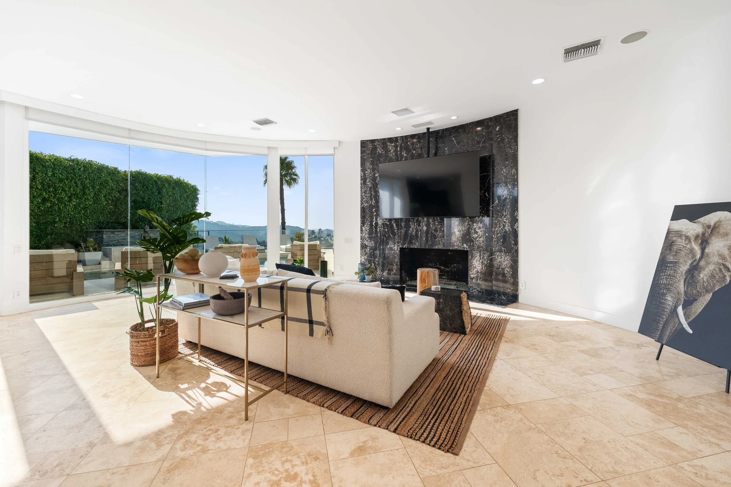Bright living room with large sliding glass doors leading to an outdoor patio, a white sofa with a blanket, potted plant, modern black marble fireplace with a mounted television, and an abstract elephant art piece leaning against the wall.