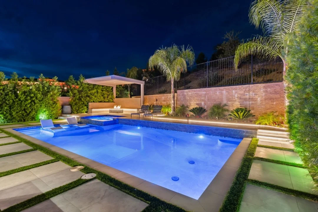 Night view of a modern backyard swimming pool with lounge chairs, palm trees, lush greenery, and outdoor seating area with lights.