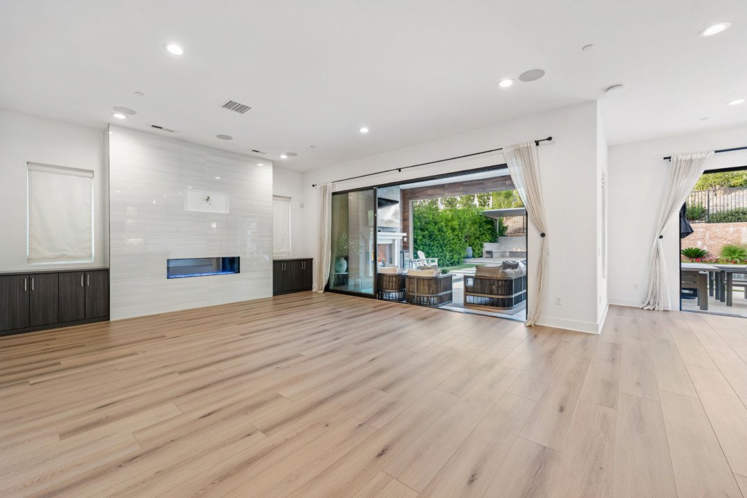 Empty living room with light wood flooring, white walls, and large sliding glass doors leading to outdoor patio with seating area.