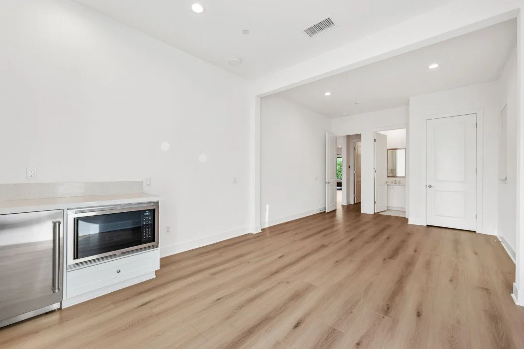 Empty room with white walls and wood flooring, featuring a stainless steel microwave and small fridge on the left, with some doors and a bathroom vanity in the background.