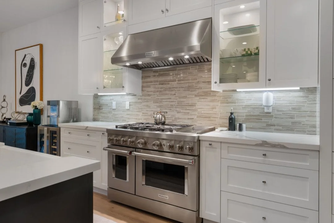 Modern kitchen with stainless steel oven and cooktop, white cabinetry, glass-front upper cabinets, a tiled backsplash, and decor accessories.