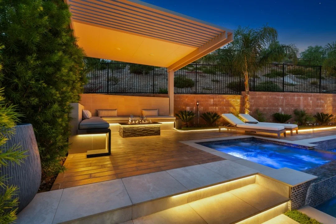 Backyard patio area at dusk with a small pool illuminated by blue lights, a lounge chair, a shaded seating area with cushions, and lush plants and trees surrounded by a brick wall.