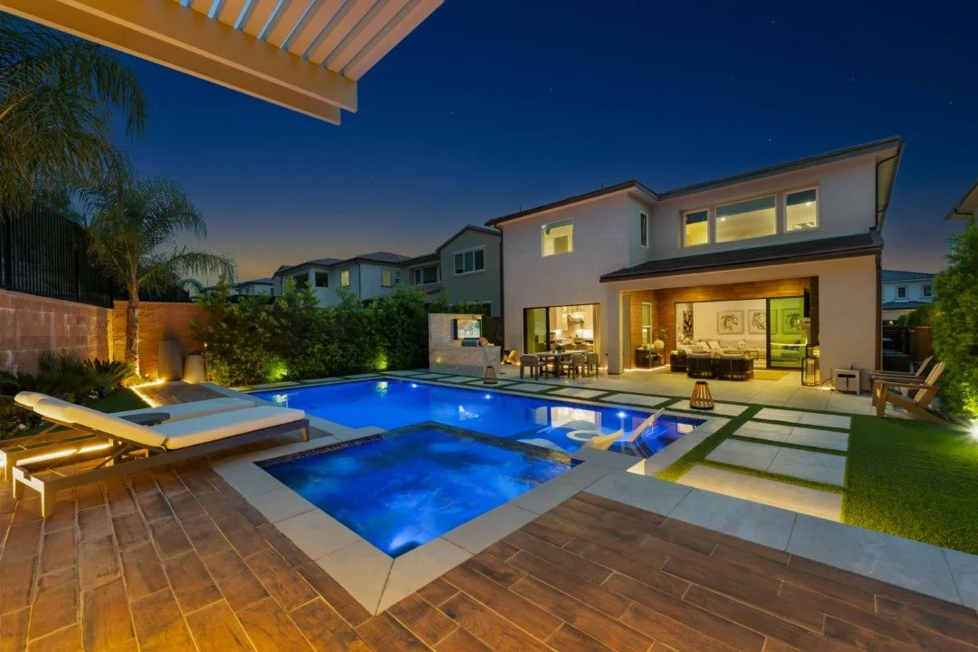 Nighttime view of a backyard with illuminated swimming pool, hot tub, outdoor lounge area, and modern multi-story house with large windows.